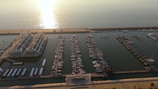 Sunset over calm marina with boats docked in rows. Water reflects golden light from setting sun. Modern piers and clear skies frame serene scene. Ideal for travel, leisure, or coastal tourism projects