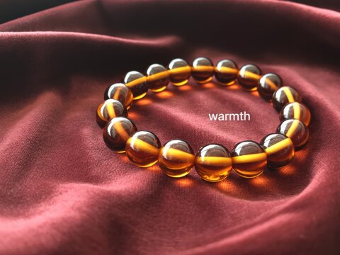 Polished amber bead bracelet on burgundy velvet fabric with glowing yellow internal bands. Circular jewelry featuring translucent spheres and centered text warmth on red cloth.