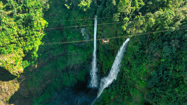 Aerial view of Tad Fane Waterfall in Laos, with a tourist enjoying tea while suspended on a zipline above the spectacular twin cascades and lush rainforest of the Bolaven Plateau.