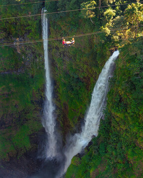 Aerial view of Tad Fane Waterfall in Laos, with a tourist enjoying tea while suspended on a zipline above the spectacular twin cascades and lush rainforest of the Bolaven Plateau.