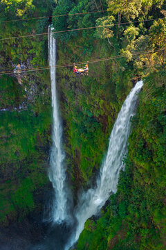 Aerial view of Tad Fane Waterfall in Laos, with a tourist enjoying tea while suspended on a zipline above the spectacular twin cascades and lush rainforest of the Bolaven Plateau.