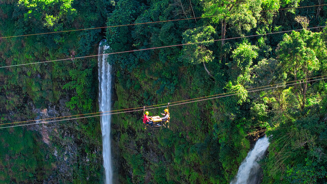 Aerial view of Tad Fane Waterfall in Laos, with a tourist enjoying tea while suspended on a zipline above the spectacular twin cascades and lush rainforest of the Bolaven Plateau.