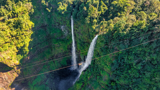 Aerial view of Tad Fane Waterfall in Laos, with a tourist enjoying tea while suspended on a zipline above the spectacular twin cascades and lush rainforest of the Bolaven Plateau.