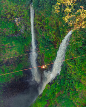 Aerial view of Tad Fane Waterfall in Laos, with a tourist enjoying tea while suspended on a zipline above the spectacular twin cascades and lush rainforest of the Bolaven Plateau.