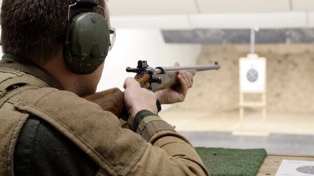 A man aims a rifle at a paper target during indoor marksmanship training at a shooting range.