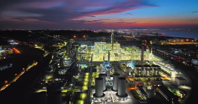 Aerial view of an illuminated chemical refinery and industrial factory plant at dusk against an evening twilight sky and coastline city lights.