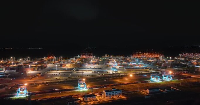 Aerial night view of a busy illuminated commercial container port terminal with gantry cranes, cargo stacks, and logistics facilities.