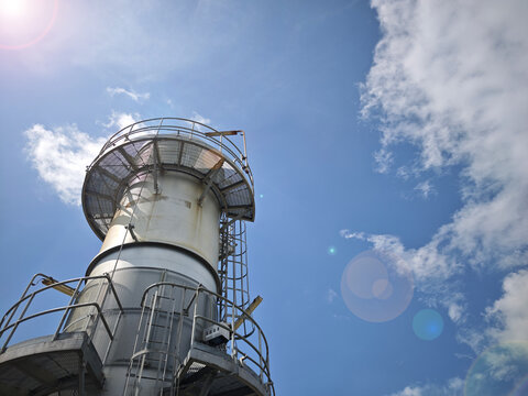 Tall industrial smokestack with metal platforms and safety ladder against a bright blue sky with sun flare. Low angle view of a factory exhaust pipe structure.