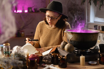 realistic witch brewing potion in cauldron. sitting at table with dried herbs and notepad. Everyday life intertwined with traditional witchcraft, magic and mystical atmosphere. © Andrii Lysenko