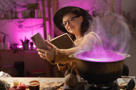 realistic witch brewing potion in cauldron. sitting at table making selfie with spells book on mobile phone. Everyday life intertwined with traditional witchcraft, magic and mystical