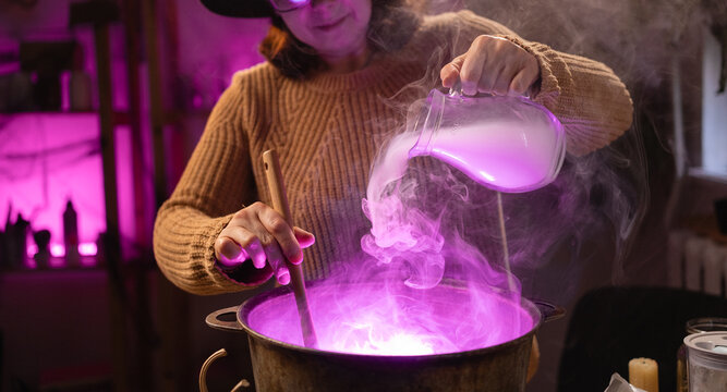 Halloween. Witch in hat brews a magic potion with smoke in cauldron on fire in scary kitchen. Fantasy.