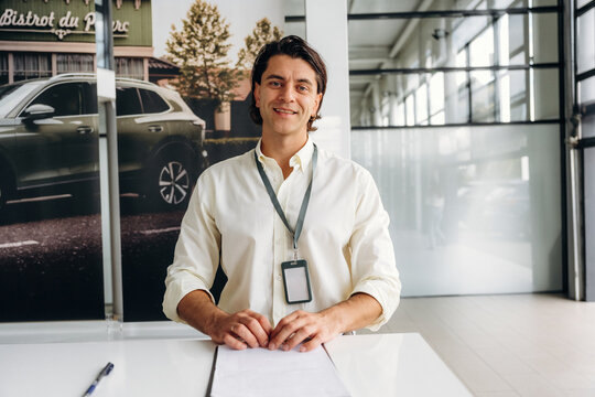 Male car dealership employee in a white shirt sits at a desk with a notepad and pen, smiling in front of a large window and promotional vehicle backdrop