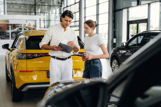 Male car sales representative in white shirt discusses vehicle features with female customer in modern auto dealership, yellow SUV prominently displayed in background
