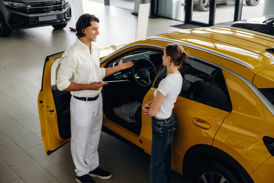Male car salesperson in white shirt engages with female customer near yellow SUV inside modern automobile showroom with large windows and vehicles on display