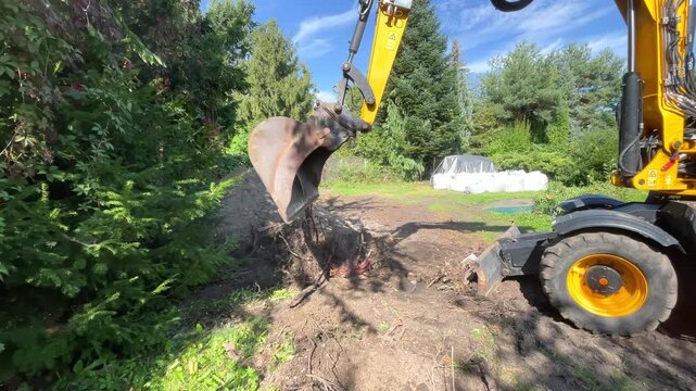 A yellow backhoe excavator works on clearing soil and rocks near a forest area outdoors, land clearing, earthmoving action, forestry service.