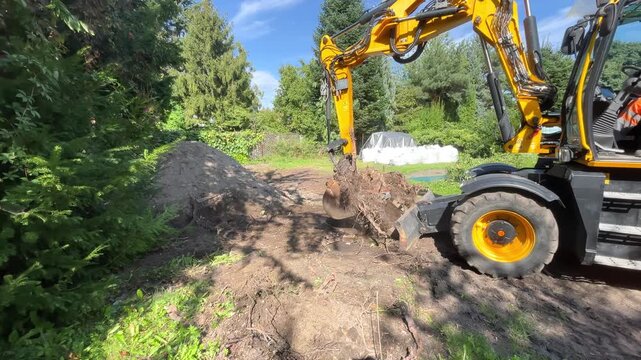 A yellow backhoe loader maneuvers through dirt ground on a construction site near green trees, land preparation, heavy machinery.