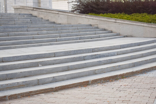 Wide exterior stone staircase made of grey granite steps leading upwards with a paved tile walkway in an urban public space during daytime