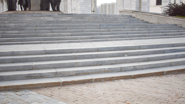 Wide exterior stone staircase made of grey granite steps leading upwards with a paved tile walkway in an urban public space during daytime