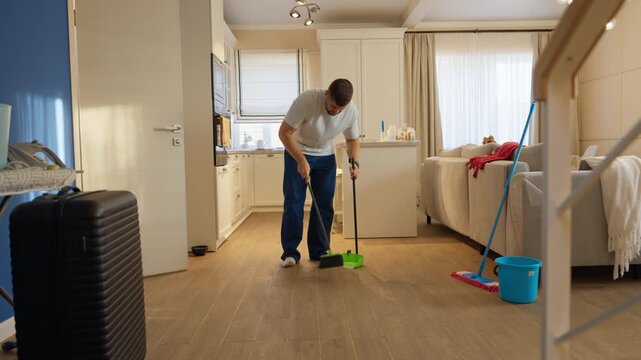 Man cleaning the living room with a broom and a mop in a bright apartment during the day