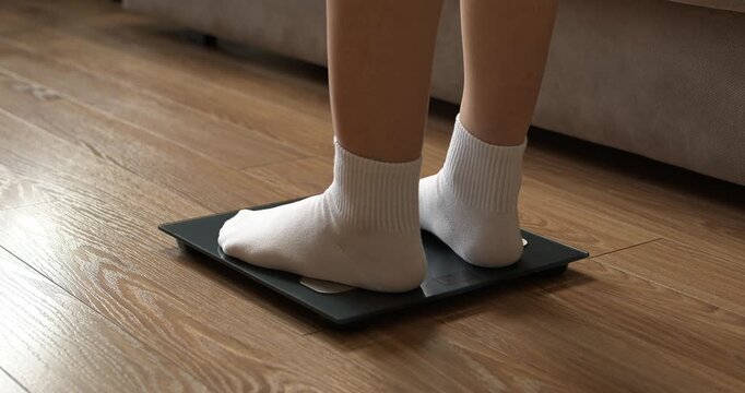Woman feet in white socks stepping on digital electronic scales to check weight on wooden floor.