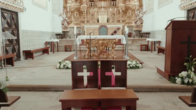 Wooden benches face a gilded baroque altar decorated with white flowers inside a tiled parish church before a wedding.