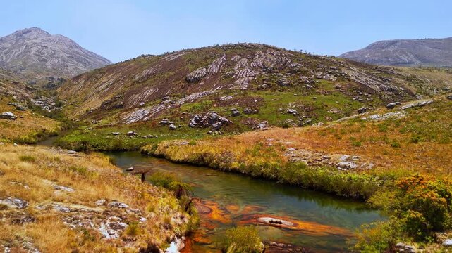 Drone glides over a winding river bordered by golden grassland and exposed rocks beneath the mountains of Mulanje Massif, Malawi.