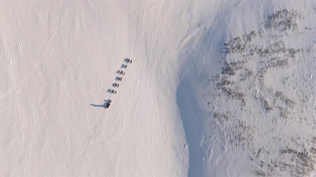 Sled dog team casting long shadows as it crosses a snowy slope in Svalbard beside a patch of exposed dark rock, orbit and ascend shot