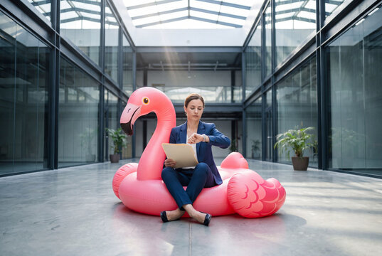Une jeune femme en costume bleu, est assise jambes crois&eacute;es sur une bou&eacute;e rose en forme de cygne avec son ordinateur, dans un hall &agrave; parois de verre au bureau, elle regarde sa montre - surr&eacute;alisme