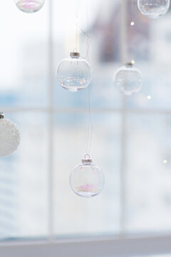 Christmas ornaments hanging against a window, delicate glass baubles with pastel decorations and sparkling lights, elegant winter holiday scene with airy festive atmosphere