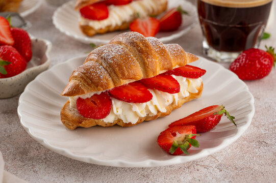 Homemade croissants filled with whipped cream and fresh strawberries on white plates with cups of coffee, milk and mint leaves. Bright breakfast or brunch scene