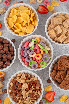 Variations of breakfast cereals in bowls. Bowl with corn pads, colorful rings, chocolate balls and cornflakes on a gray concrete background. Top view, copy space.