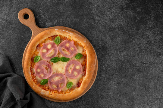 Halloween pizza with smiling ham faces, basil leaves and spooky decorations on a dark background, creative seasonal food concept for parties, menus, and social media. Top view. Copy space.