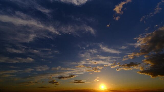 Time lapse of a tranquil sunset with orange glow on dark blue sky and illuminated moving clouds. A calm nature scenery with the glowing sun and good color contrast. 
