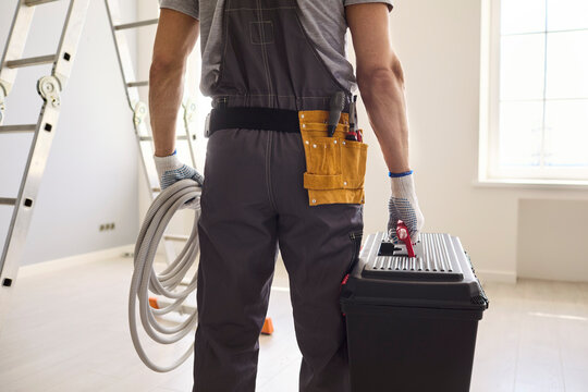 Electrician worker holds toolbox and cable indoors. Rear view of technician beside ladder, prepared for home installation and renovation. Reliable residential maintenance service concept.