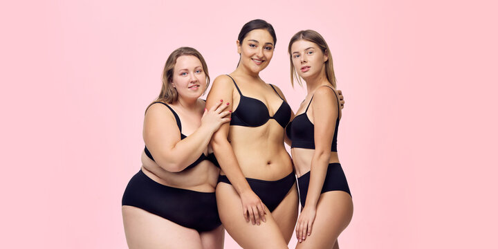 Trio of confident women stands proudly in black underwear in pink studio. Diverse beauty, lingerie advertising, self acceptance concept.