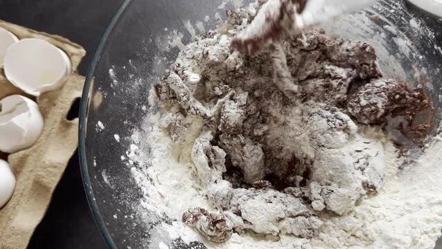 Adding and mixing flour to chocolate dough in a glass bowl, showing a hand using a spatula