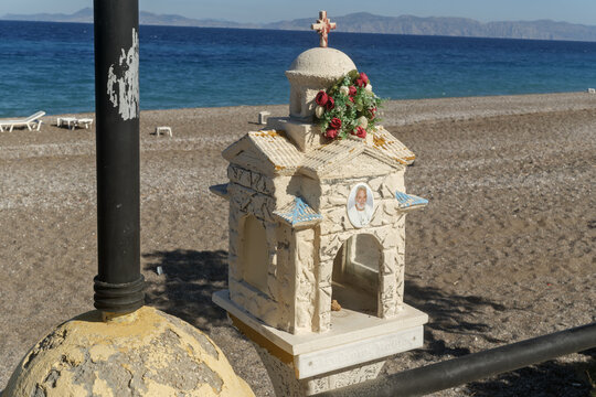 A small roadside shrine with a dome and cross stands by the Rhodes shore, its flowers and portrait glowing in clear sun beside calm blue water.