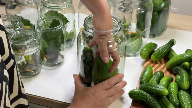 a person puts green cucumbers into a glass jar, several other jars with leaves and water stand on a white table, a wooden board holds more cucumbers