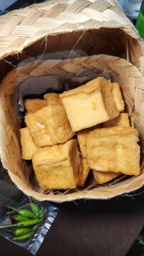 Crispy fried tofu cubes are arranged in a woven bamboo basket, highlighting Indonesian cuisine. The golden-brown tofu sits on a paper lining, showcasing its textured surface. Adjacent to the basket, f