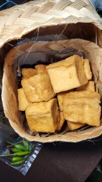 Crispy fried tofu cubes are arranged in a woven bamboo basket, highlighting Indonesian cuisine. The golden-brown tofu sits on a paper lining, showcasing its textured surface. Adjacent to the basket, f