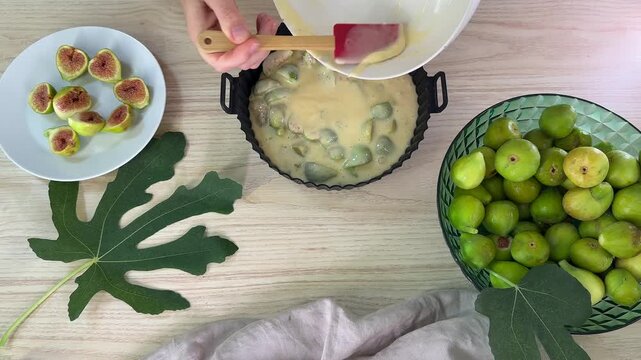 A person pours batter from a white bowl into a fluted cake pan containing fruit, with a plate of sliced figs and a bowl of whole green figs nearby
