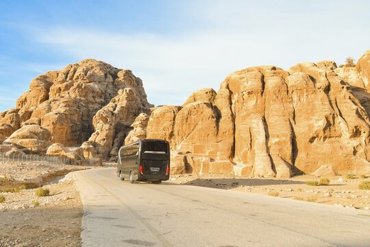 Black passenger coach bus driving asphalt road winding through sandstone rock mountains canyon desert landscape leading ancient archaeological tourist attraction Little Petra Jordan