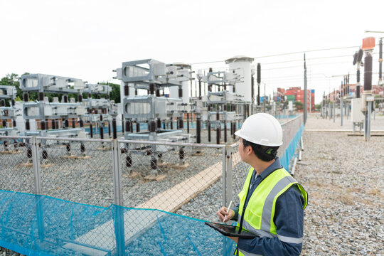 Inspecting capacitor equipment during a substation inspection, a technician examines the capacitor bank with a tablet beside a lattice tower.