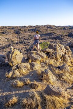 Young woman on rocks, barren landscape with rocky hills and acacia trees, African savannah in the evening light, Hobatere Concession, Kunene, Namibia