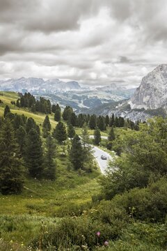 Aerial cinematic view car vehicle drives Winding mountain road through Dolomites landscape with alpine forest, rustic cabins, green meadows and dramatic peaks, ideal road trip travel concept