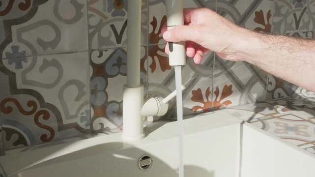Pulling head from pulldown kitchen faucet to rinse white sink with water spray. Sunlit patterned ceramic tiles backdrop. Home interior and lifestyle themes.