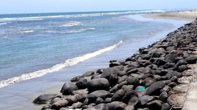 A rocky breakwater embankment made of dark boulders lining a sandy beach and blue ocean surf.