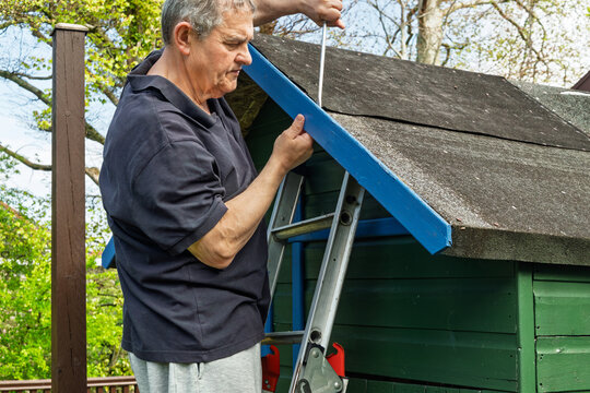 Older man on stepladder removes blue-painted timber fascia board from felt roof of small green wooden garden playhouse, screwdriver in hand, trees visible behind