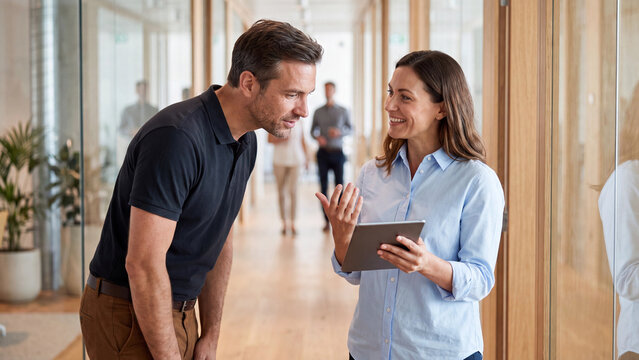 Office workers interact in a wooden corridor as a man leans down to view a presentation on a digital tablet held by his colleague.