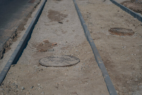 Unpaved sidewalk reconstruction process with concrete curb stone borders, manhole covers, and prepared gravel foundation layer.
